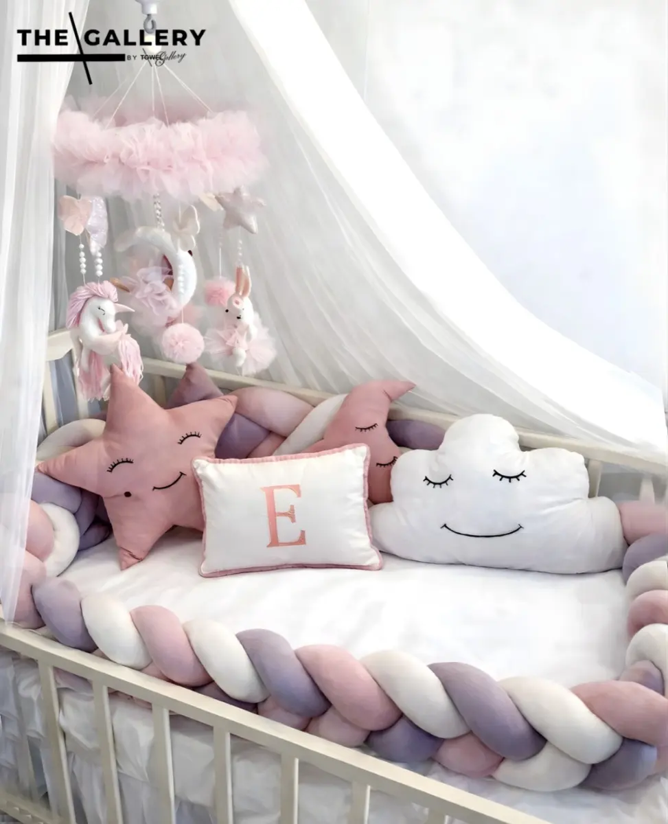 A cot styled with The Gallery's personalised newborn set — braided bumper, star and cloud cushions, tulle canopy.